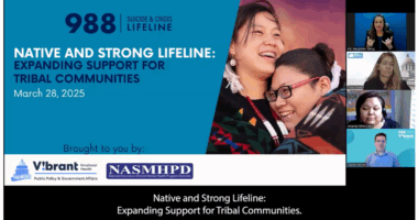 A webinar slide titled “Native and Strong Lifeline: Expanding Support for Tribal Communities – March 28, 2025” appears on the left, featuring the 988 Suicide & Crisis Lifeline branding and logos for Vibrant Emotional Health and NASMHPD. The slide includes a photo of two Native youth wrapped in a colorful blanket, smiling and embracing. On the right side of the screen, four video panels show an ASL interpreter, a speaker with a state capitol background, another speaker, and Daniel Herron in a Vibrant-branded virtual background. The caption bar below reads: “Native and Strong Lifeline: Expanding Support for Tribal Communities.”